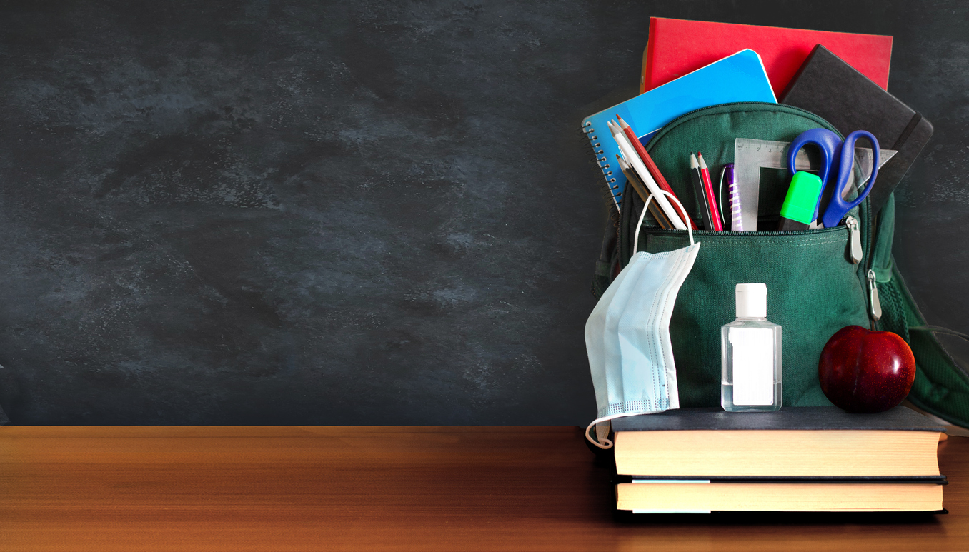 school back and a stack of books sitting on a desk in front of a blackboard with a bottle of hand sanitizer showing how covid has impacted education