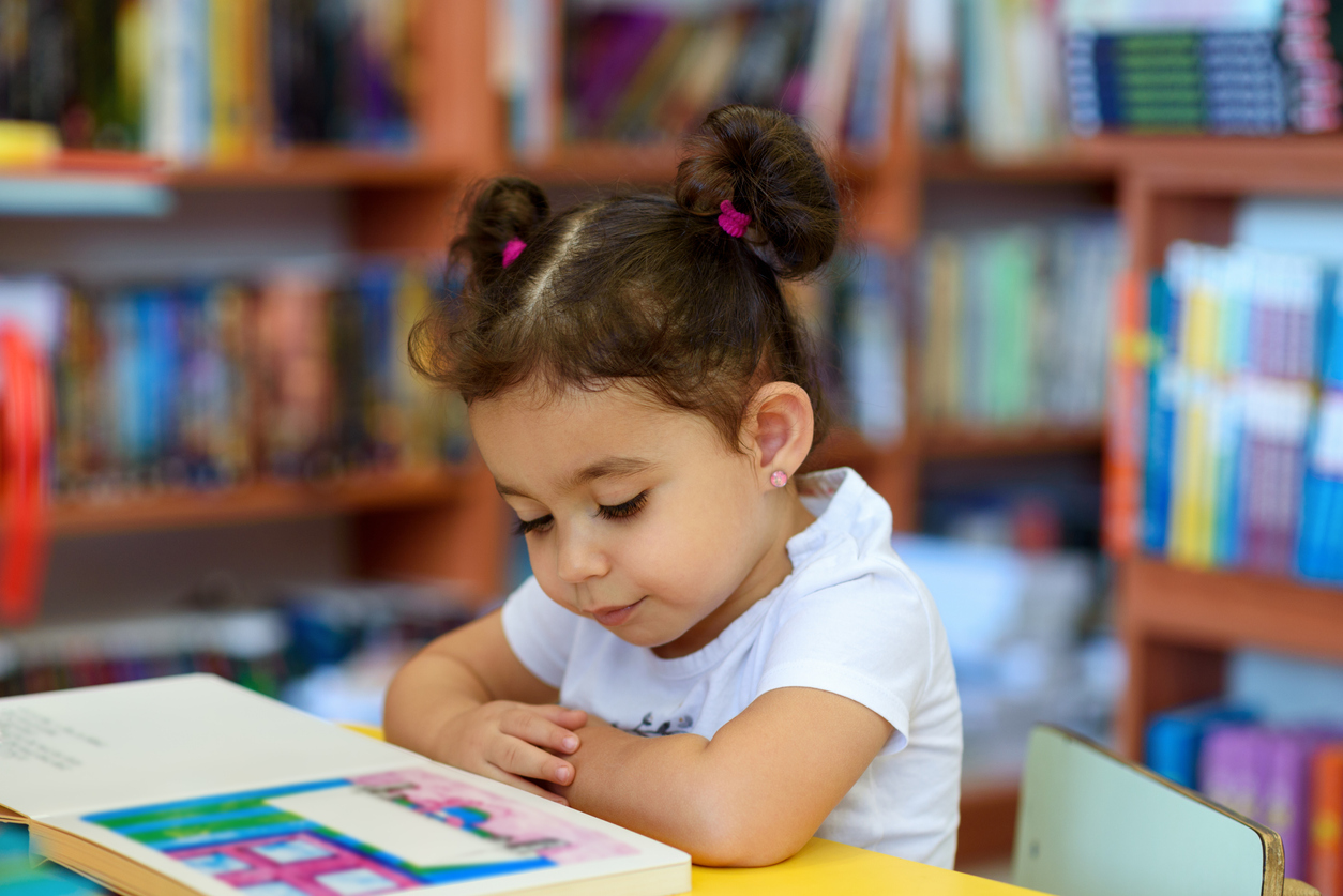 Little Girl Indoors In Front Of Books. Cute Young Toddler Sitting On A Chair Near Table and Reading Book. Child reads in a bookstore, surrounded by colorful books. Library, Shop, Shelving In Home.