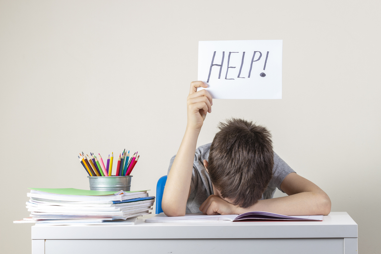A tired, frustrated boy with dyslexia sits at a table with his head down on his arm, holding up a “Help!” sign